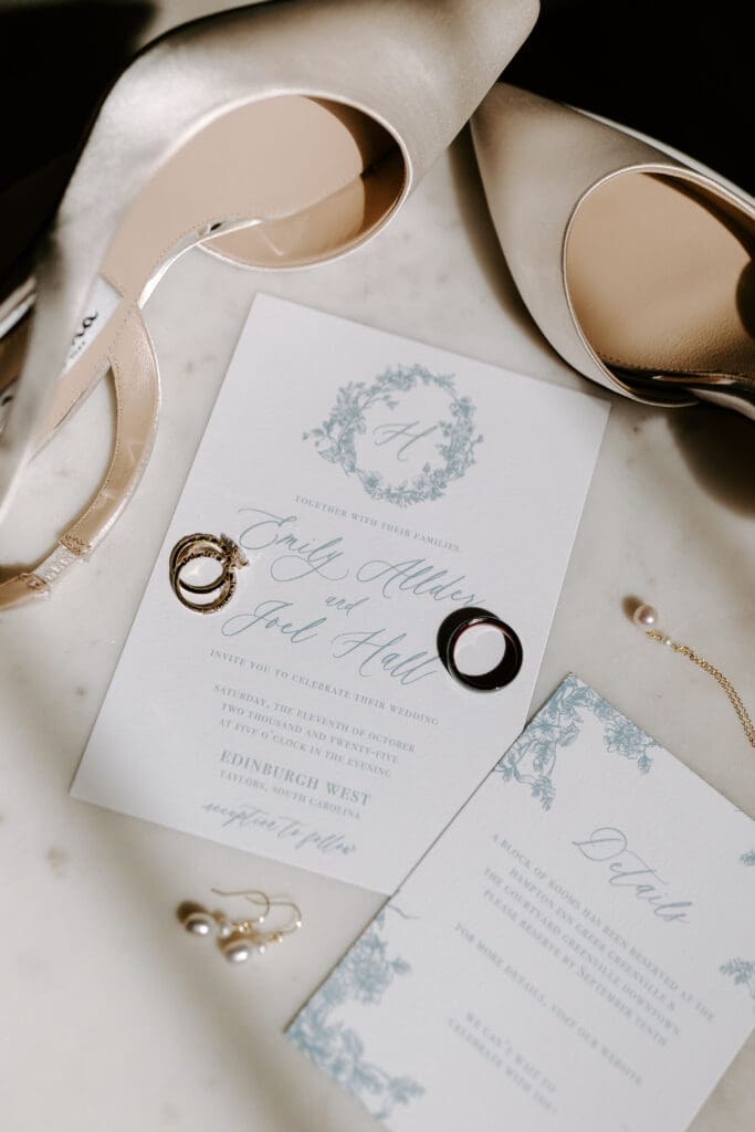 Image of Wedding Invitation and Details card in dusty blue ink with floral decoration sitting on top of a marble table surrounded by wedding shoes, wedding rings, and earrings.