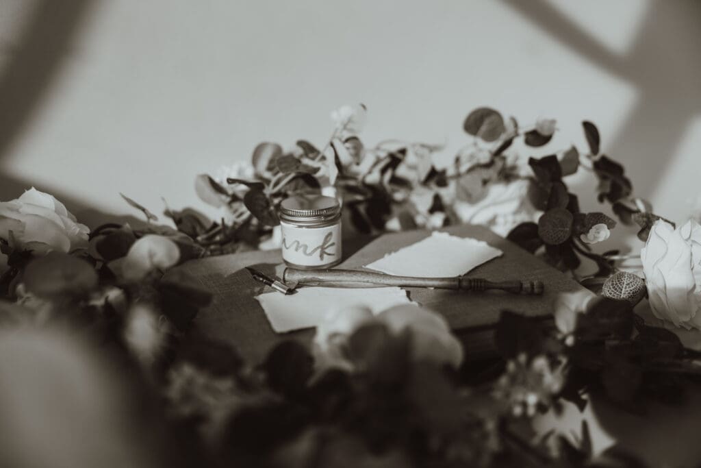 Black and White Image of a Oblique Calligraphy Pen sitting on top of a book with raw paper place cards and a jar of ink surrounded by florals.