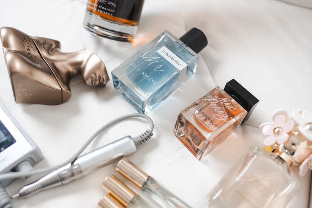 Image of Multiple bottles of engraved perfume and cologne sitting on a fabric background with a handheld engraving device sitting next to them.